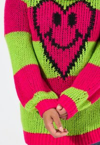 Pull en tricot avec des rayures rose vif et vert, coupe oversized, et un grand motif de cœur souriant noir à l'avant. La texture est lâche et aérienne.