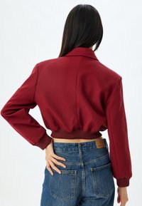 Afskåret rød jakke med krave, ribbede manchetter og kant. Stoffet er glat, og den er parret med højtaljede blå denim jeans.