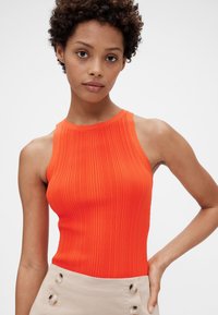 Ribbet orange ærmeløs top med høj hals, kombineret med et beige skørt med store knapper på siden. Glat, tætsiddende tekstur.