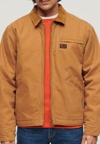 Bruine katoenen jas met corduroy kraag, ritssluiting aan de voorkant en borstzak, gedragen over een oranje trui, gecombineerd met blauwe jeans.