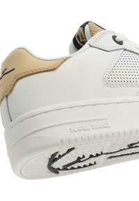Zapatilla blanca de cuero con acentos en tono marrón, detalles perforados y suela texturizada. Presenta un diseño de caña baja y un logo distintivo en el talón.
