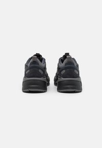 Un paio di sneakers Puma RS-X grigio scuro e nero viste da dietro, che evidenziano le suole spesse e i ganci sul tallone su uno sfondo bianco.