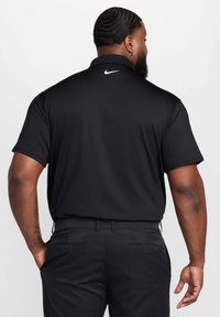 Czarna koszulka polo Nike z kołnierzykiem, krótkimi rękawami, gładką fakturą oraz białym logo na górnej części pleców. Noszona z czarnymi spodniami.