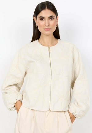 Vrouw met lang donker haar draagt een crèmekleurige geborduurde bloemenritsjas en bijpassende broek, handen in de zakken tegen een witte achtergrond.
