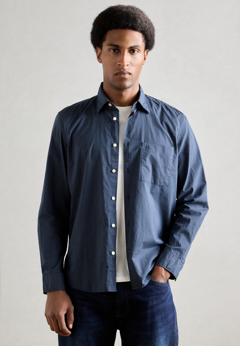 Ναυτίες μπλε καρό πουκάμισο με κουμπιά και τσέπη στο στήθος, συνδυασμένο με άσπρη μπλούζα με στρογγυλή λαιμόκοψη, φορεμένο με σκουρόχρωμο τζιν.