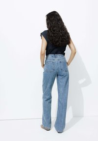 Jean bleu taille haute avec une coupe ample, en denim. Doté de poches arrière et d'une coupe droite. Porté avec un haut noir, de dos.
