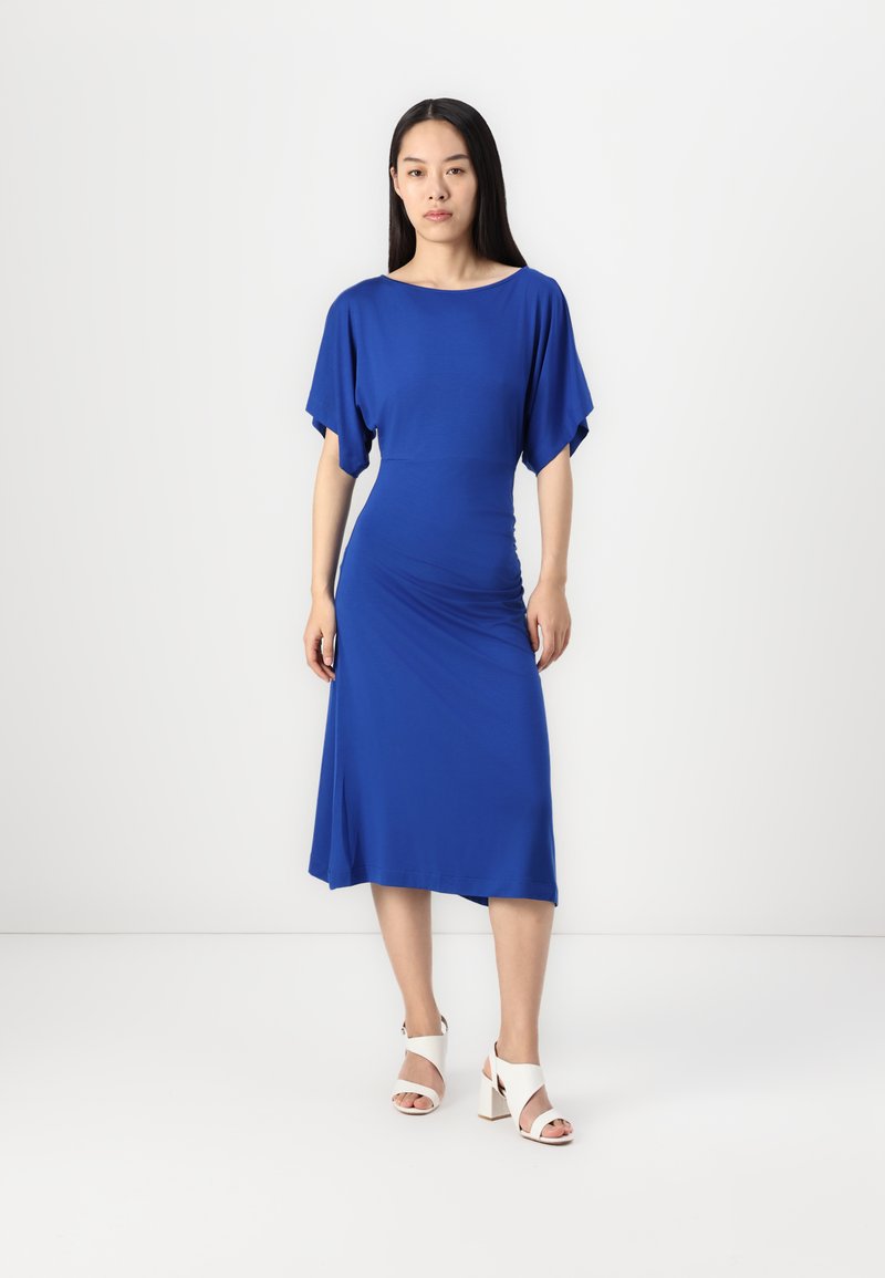 Blaue Midi-Kleid mit Fledermausärmeln, figurnahem Oberteil, asymmetrischem Saum, kombiniert mit weißen Blockabsatzsandalen.