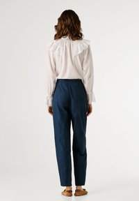 Λευκό μπλουζάκι με βολάν στους ώμους, συνδυασμένο με σκούρο μπλε παντελόνι. Το μοντέλο φοράει καφέ παπούτσια τύπου slip-on. Απλός, καθαρός σχεδιασμός.