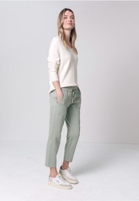 Kobieta stojąca z rękami w kieszeniach, ubrana w biały sweter, szałwiowozielone spodnie i białe trampki na jednolitym szarym tle.