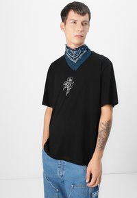 Sort bomulds T-shirt med korte ærmer, der har hvid blomsterbroderi. Modellen har et blåt paisley-halstørklæde om halsen.