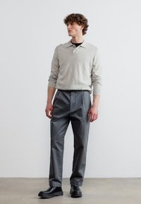 Νεαρός άνδρας στέκεται φορώντας ανοιχτό γκρι πόλο με μακρύ μανίκι, σκούρα γκρι πλισέ παντελόνια και μαύρα παπούτσια, μπροστά σε ένα απλό λευκό φόντο.