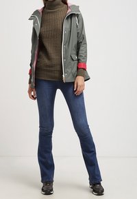 Chaqueta verde impermeable con interior rosa, combinada con un suéter texturizado en oliva y unos jeans de campana azul oscuro. Zapatillas negras completan el conjunto.