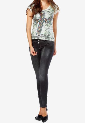 Femme portant un T-shirt blanc à imprimé floral en croix, un jean skinny foncé et des chaussures à talons noires, debout avec une main près de la poche.