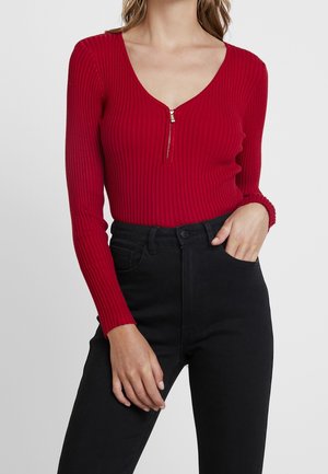 Frau trägt ein rotes, geripptes Langarmshirt mit einem vorderen Reißverschluss und schwarze, hoch taillierte Hosen, eine Hand in der Tasche, vor einem schlichten Hintergrund.