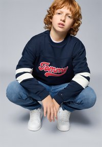 Marineblaues Sweatshirt mit rotem "Tommy"-Logo, weißen Streifen an den Ärmeln, kombiniert mit blauen Jeans und weißen Sneakers. Weiche Textur, lässiges Design.