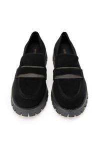 Scarpe slip-on in suede nero con due accenti di cinturini metallici e una suola in gomma nera spessa e testurizzata. Punta arrotondata e design a profilo basso.