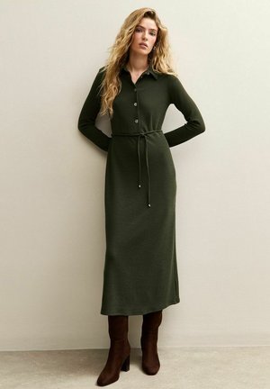Kobieta z długimi falującymi włosami, ubrana w ciemnozieloną, zapinaną na guziki sukienkę midi z paskiem w talii oraz brązowe botki na obcasie, stojąca na tle jasnej ściany.