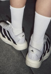 Personne portant des baskets blanches Adidas avec des bandes noires et des chaussettes blanches côtelées, debout sur des carreaux de sol gris.