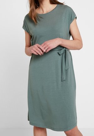 Vrouw draagt een knie-lange jurk in saliegroen met korte mouwen en een strik aan de zijkant van de taille, staat met de handen voor zich geklapt.