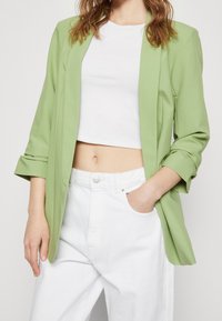 Grön blazer i ett mjukt tyg, med krage, trekvartsärmar och sidfickor, parat med en vit kort topp och vita byxor.