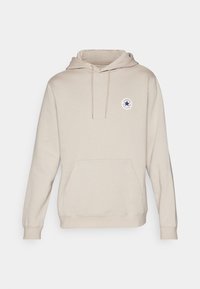 Beige hoodie med en kenguruficka, dragsko i huvan och en rund logopatch med en blå stjärna på bröstet. Mjuk tygtextur.