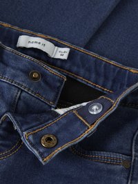 Tummansiniset farkut, joissa on oranssit kontrastitikkaukset. Vyötäröllä on merkki ja metallinen nappisuljin sekä muovinen nappi.