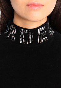 Svart polotröja i mjukt tyg med ribbat krage. Kragen har glittriga rhinestone-bokstäver som stavar "R" och "D."