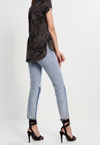 Šedý maskáčový top s krátkým rukávem a bočními rozparky, doplněný o světle modré potrhané džíny se tmavě modrým bočním panelem a černými sandály na podpatku.