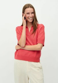 Koralle Kurzarm-Polohemd mit V-Ausschnitt, gerippten Bündchen und Saum, kombiniert mit hellbeigen Leinenhosen. Glatte Stricktextur.