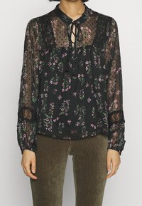 Blusa nera trasparente decorata con piccoli motivi floreali e pois, con dettagli in pizzo e un collo con lacci. Maniche lunghe con polsini elasticizzati.