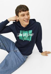 Donkerblauwe hoodie met een grafisch ontwerp aan de voorkant in groen en wit. Heeft een kangoeroezak en een capuchon met trekkoord. Gecombineerd met lichtblauwe jeans.