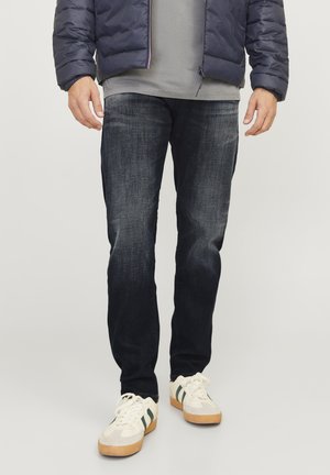 Homme portant un jean foncé, des baskets blanches avec des rayures vertes, une chemise grise et une doudoune bleu marine, debout sur un fond uni.