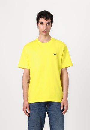 T-shirt in cotone gialla con maniche corte, colletto rotondo e un piccolo logo Lacoste sul lato sinistro del petto, abbinata a jeans blu.