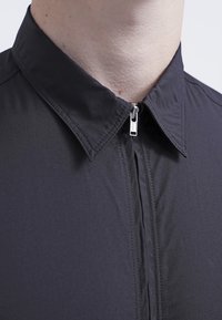 Chemise noire à fermeture éclair avec col pointu, texture lisse et détails de couture subtils le long de la ligne de la fermeture éclair.