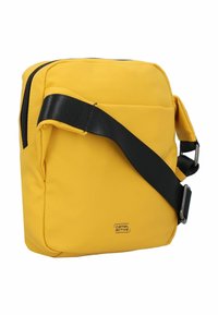 Gelbe Umhängetasche aus strukturiertem Nylon. Sie verfügt über einen schwarzen verstellbaren Riemen, einen Reißverschluss und ein Markenlogo auf der Vorderseite.