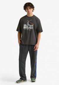 T-shirt grigia oversize con stampa grafica, abbinata a pantaloni scuri con una striscia laterale blu e scarpe da ginnastica verdi.