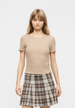 Jeune femme portant un haut beige texturé à manches courtes et une jupe plissée à carreaux marron, blanc et noir, debout devant un fond uni.