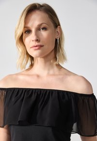 Rochie neagră, fără umeri, cu un guler din volane transparente, corset ajustat și textură netedă; asortată cu cercei lungi, strălucitori.