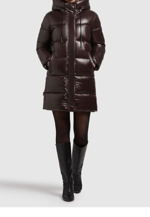 Femme portant un manteau matelassé brillant brun foncé longueur genou avec capuche, des collants noirs, et des bottes en cuir noires montant jusqu'aux genoux, debout devant un fond uni.