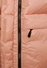 Peach puffer jacket with a black zipper, featuring vertical quilting, velcro accents, and a fabric loop detail near the zipper.