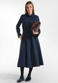Abito blu navy con bottoni, vita con cravatta, maniche lunghe e gonna fluente. Porta una borsa a tracolla trapuntata marrone. Collant neri e scarpe basse blu.