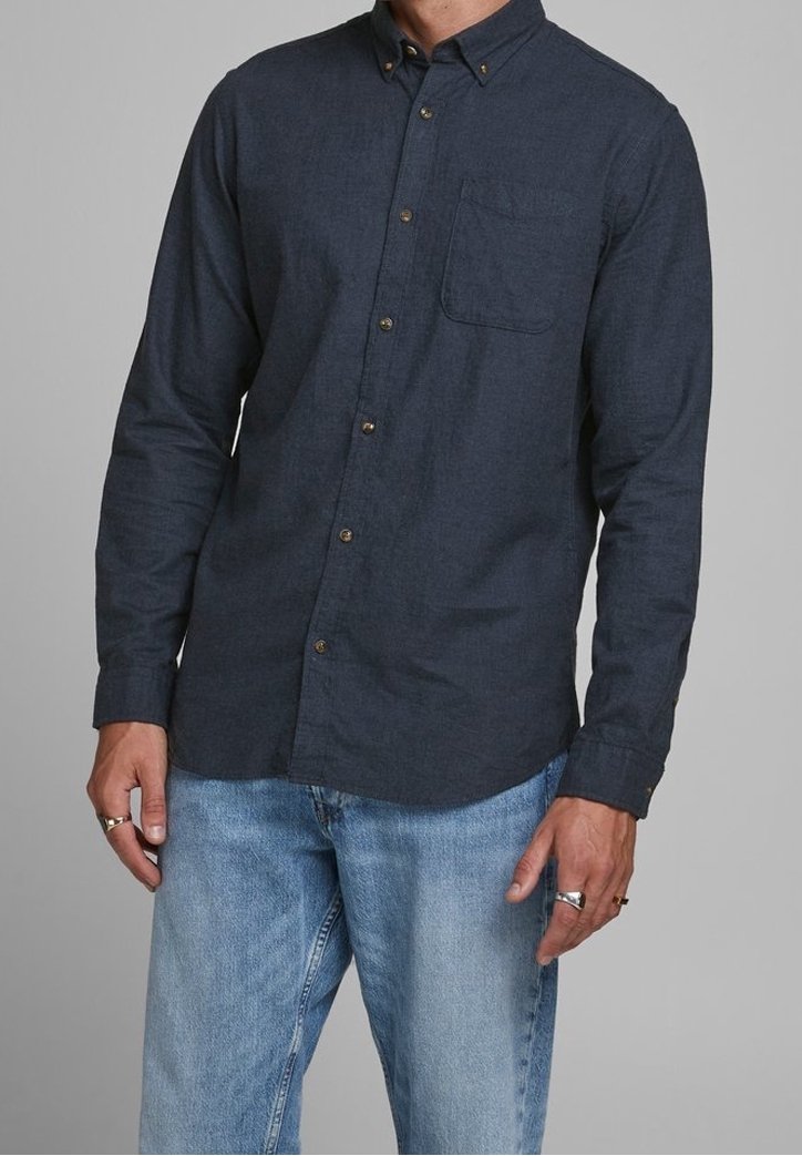 Camisa azul oscuro abotonada con tejido texturizado, que presenta un bolsillo en el pecho, cuello abotonado y botones de tono latón. Combinada con vaqueros de mezclilla claros.