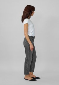 Žena stojaca v profile oblečená v bielej blúzke s krátkym rukávom, vysokými čiernymi džínsami a čiernymi špicatými balerínkami na pozadí sivošedej farby.