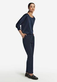 Granatowa satynowa bluzka z szerokim dekoltem i rękawami o długości trzy czwarte, zestawiona z dopasowanymi granatowymi spodniami i czarnymi loafersami.