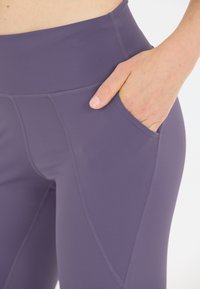 Närbild av hand i fickan på lila hight-waisted leggings med synliga sömmar mot vit bakgrund.