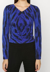 Modrý dlouhý rukáv s výstřihem do V a černým abstraktním vzorem štětcem, s nařaseným centrálním detailem, spárovaný s texturovanými černými kalhotami.