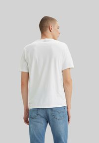 Hombre de espaldas vestido con una camiseta blanca de manga corta y jeans de denim azul claro frente a un fondo gris.