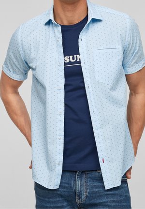 Camicia azzurra a maniche corte con frontale a bottoni, caratterizzata da un piccolo motivo, taschino sul petto e indossata sopra una maglietta blu navy.