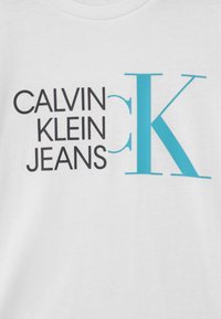 Camiseta blanca de algodón con texto negro audaz "CALVIN KLEIN JEANS" y un gran acento turquesa "K", cuello redondo y textura suave.