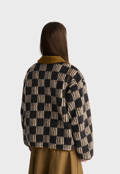 Femme aux longs cheveux bruns porte une veste en polaire à carreaux noirs et beiges avec un col moutarde, debout devant un fond gris uni.